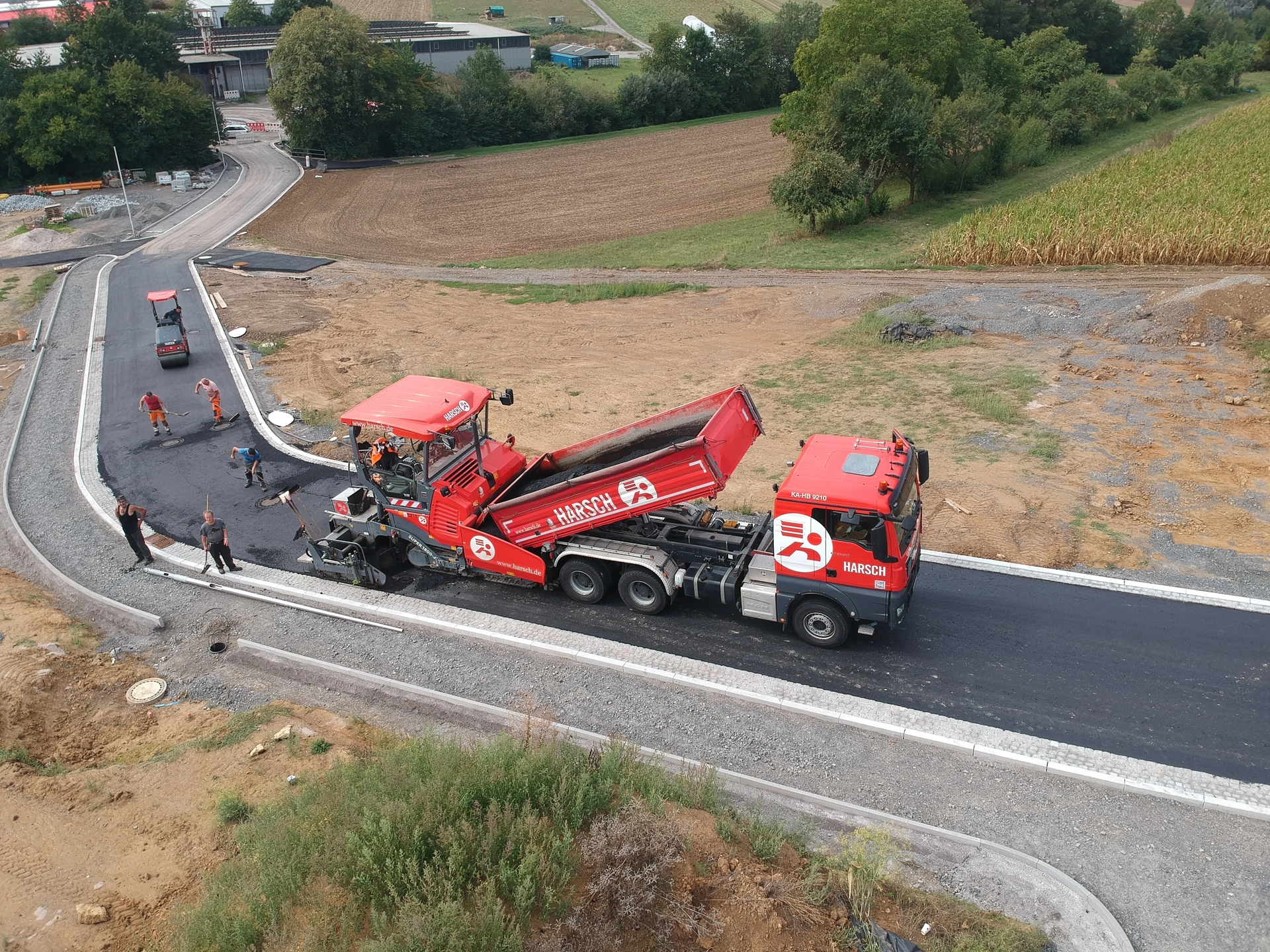 Asphaltarbeiten im Straßenbau mit Fertiger, Lkw und Baustellenpersonal aus der Luftperspektive.