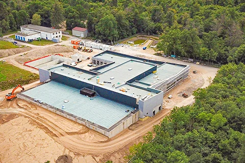 Luftaufnahme eines Gebäudes im Bau mit Flachdach, Baustellenfahrzeugen, Erdarbeiten und umliegendem Waldgebiet