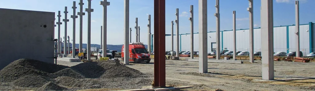 Industriebaustelle mit aufgestellten Betonfertigteilstützen, Schotterflächen, Fundamentarbeiten und Baustellenfahrzeugen