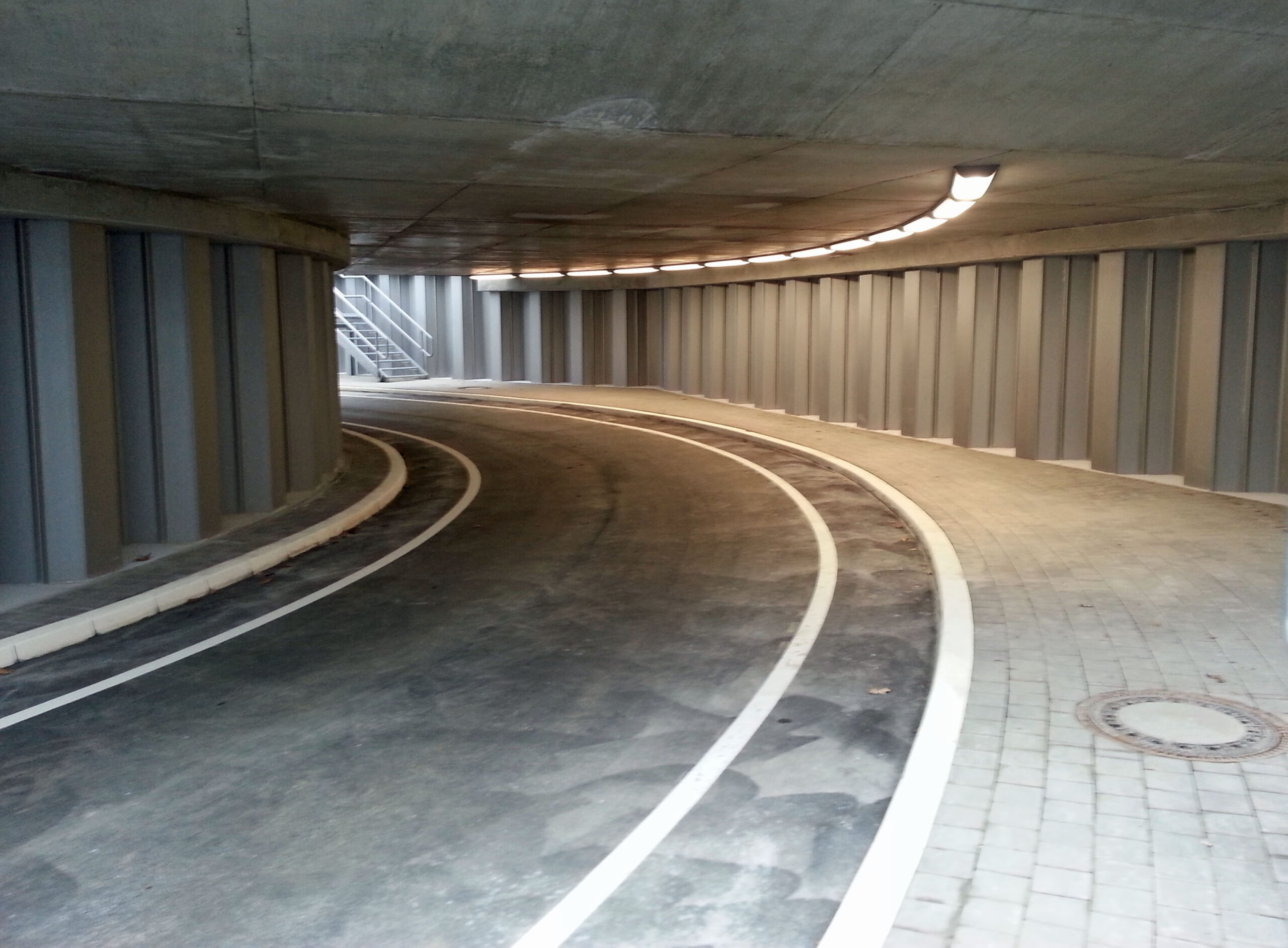 Unterirdische Zufahrtsrampe mit gebogener Fahrbahn, Betonwänden und Beleuchtung.